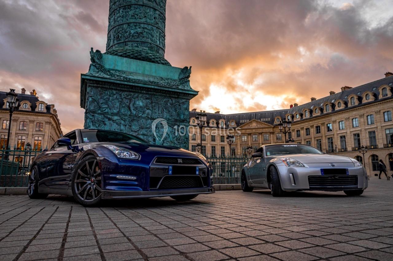 Deux voitures sportives garées sur une place pavée devant une colonne et des bâtiments historiques au coucher du soleil.