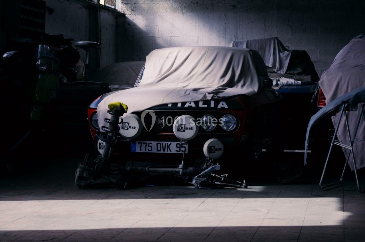 Voiture ancienne partiellement couverte dans un garage sombre, entourée de pièces détachées et d'ombre.