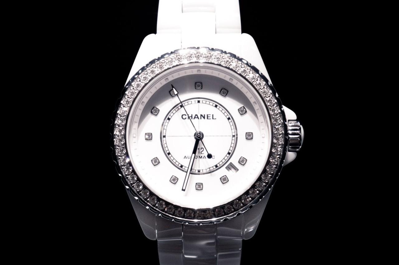 Montre blanche avec cadran rond, index en diamants et lunette sertie de pierres brillantes sur fond noir.