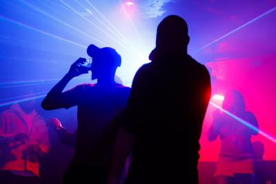 Femme assise sur un banc en extérieur, portant un sweat noir, avec un ciel bleu et des bâtiments en arrière-plan.