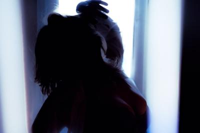 Femme assise sur un banc en extérieur, portant un sweat noir, avec un ciel bleu et des bâtiments en arrière-plan.