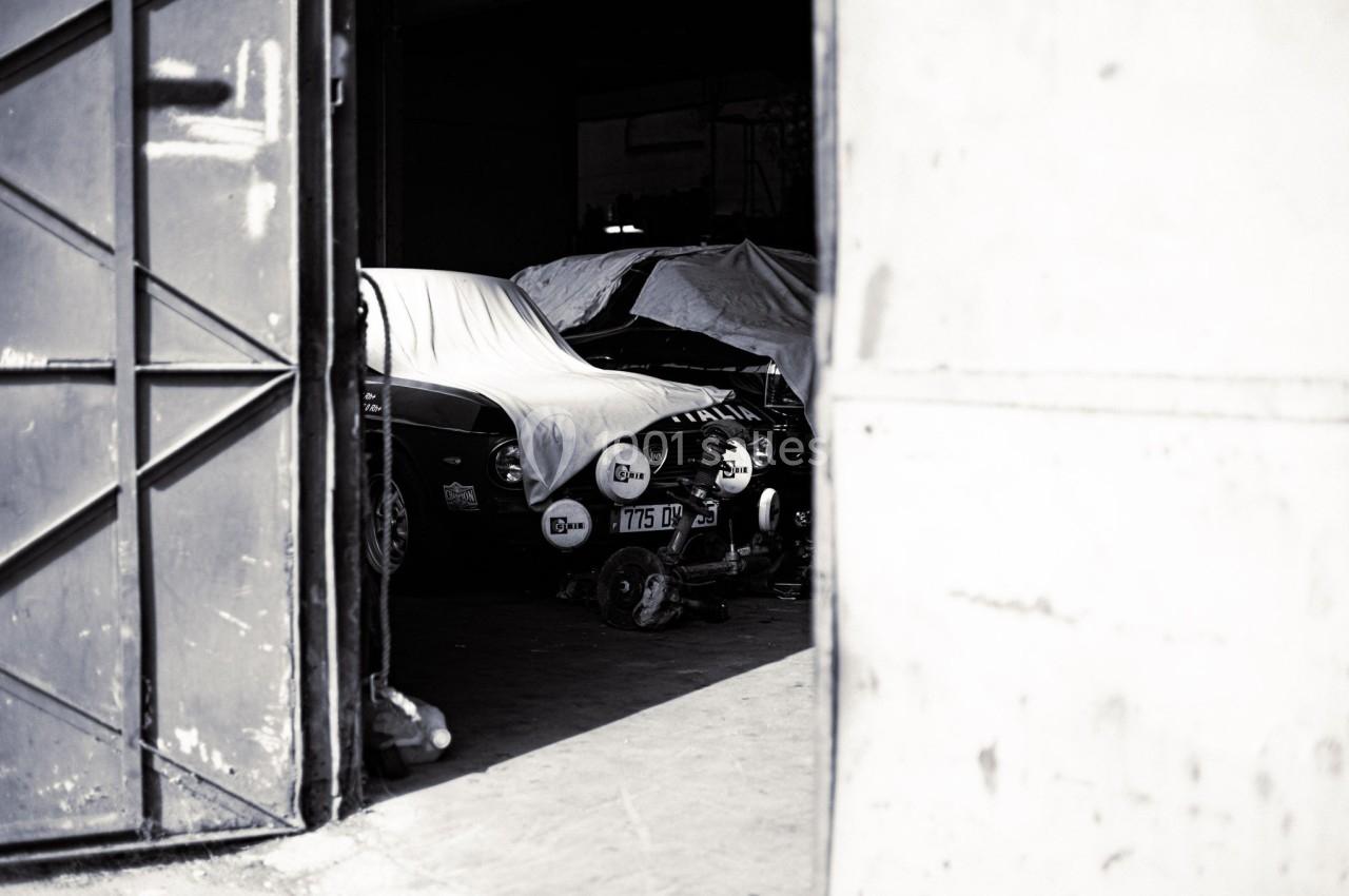 Voitures anciennes partiellement couvertes sous une bâche dans un garage sombre, vue à travers une porte entrouverte.