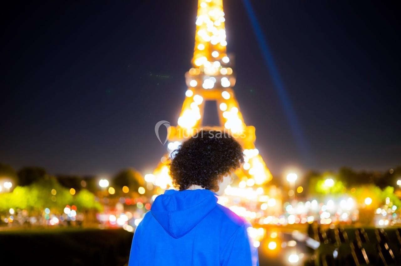 Une personne de dos en sweat bleu regarde la Tour Eiffel illuminée de nuit, entourée de lumières urbaines.