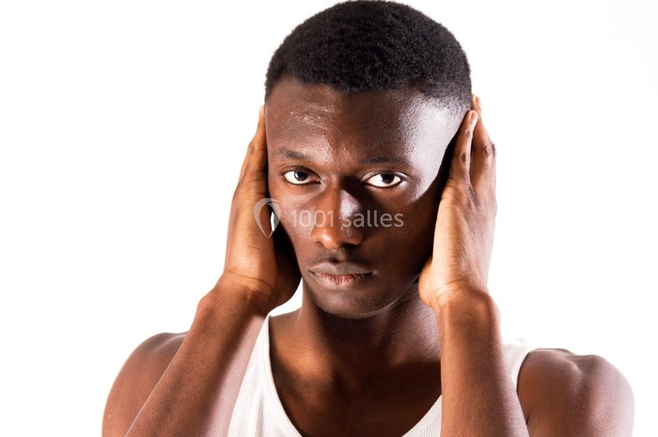 Un homme en débardeur blanc se tient debout, les mains couvrant ses oreilles, sur un fond blanc.