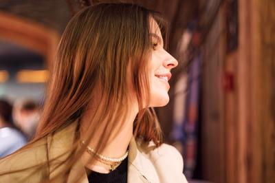Femme assise sur un banc en extérieur, portant un sweat noir, avec un ciel bleu et des bâtiments en arrière-plan.