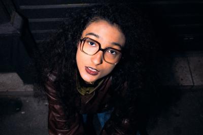 Femme assise sur un banc en extérieur, portant un sweat noir, avec un ciel bleu et des bâtiments en arrière-plan.