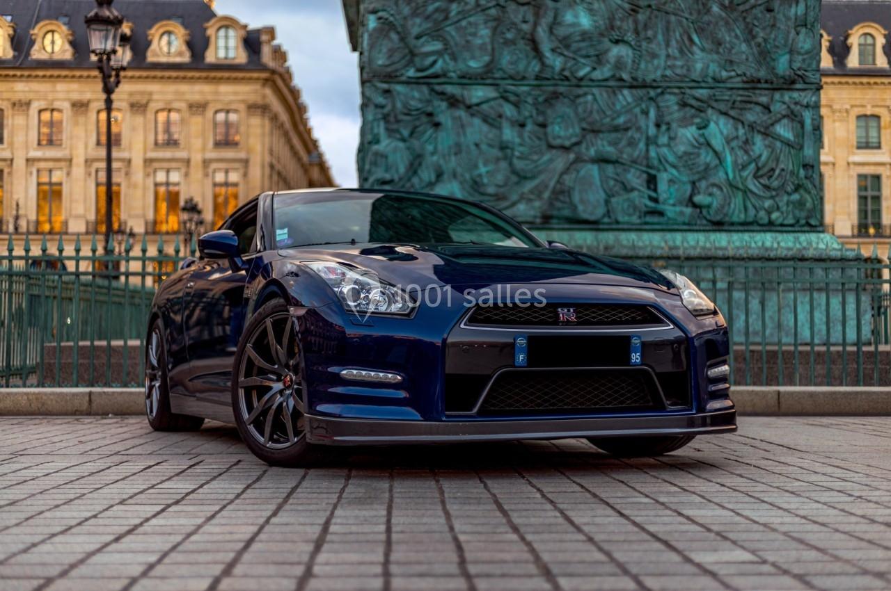 Voiture sportive bleue garée sur une place pavée devant des bâtiments historiques et une colonne monumentale.
