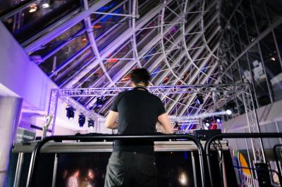 Femme assise sur un banc en extérieur, portant un sweat noir, avec un ciel bleu et des bâtiments en arrière-plan.