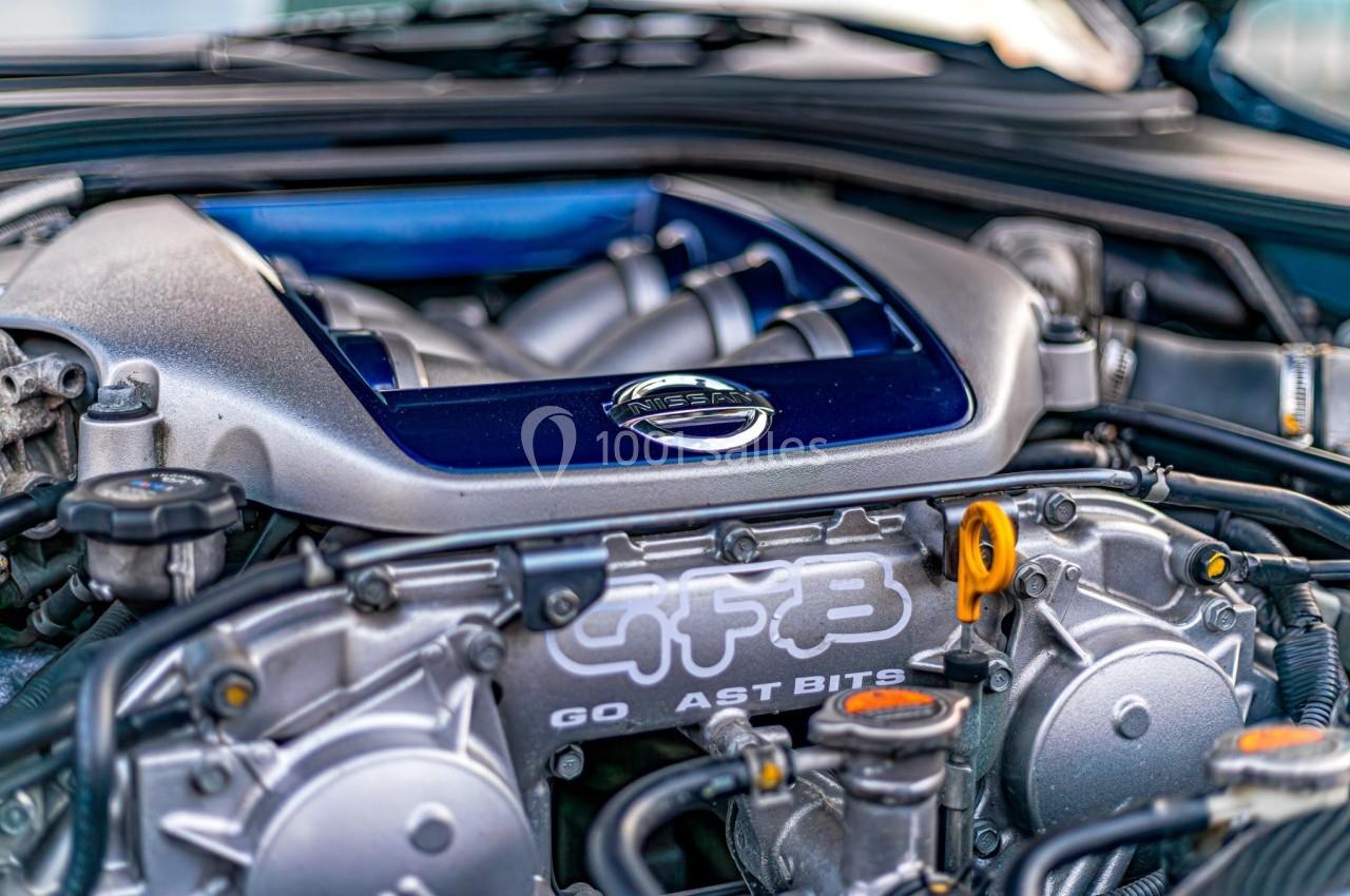 Moteur de voiture avec logo Nissan visible, entouré de composants mécaniques et détails métalliques.