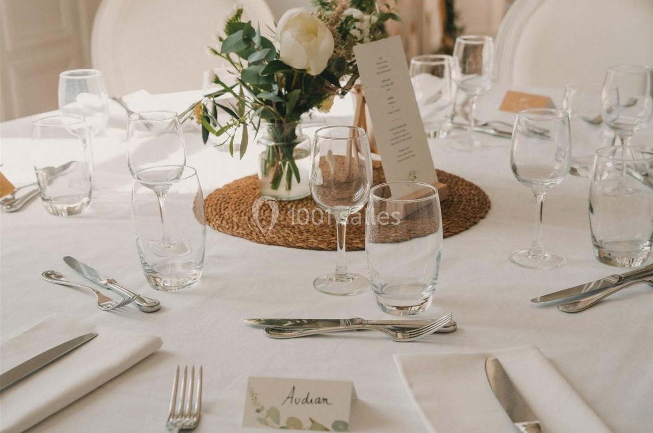 Table dressée avec nappe blanche, couverts, verres, centre de table floral et marque-place personnalisé.