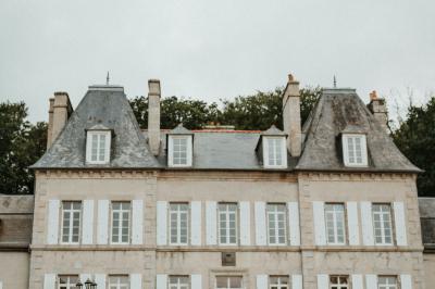 Un couple de mariés marche vers un château en pierre avec des volets blancs, entouré de quelques invités.