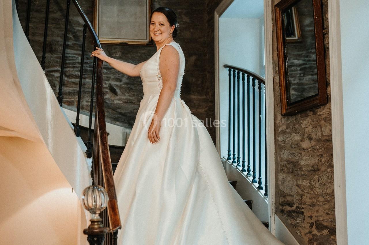 Une femme en robe de mariée blanche pose sur un escalier en bois dans un intérieur aux murs en pierre.
