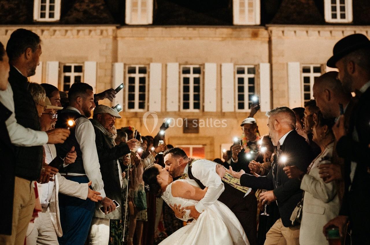 Un couple de mariés s'embrasse sous les applaudissements des invités, devant un bâtiment éclairé de nuit.