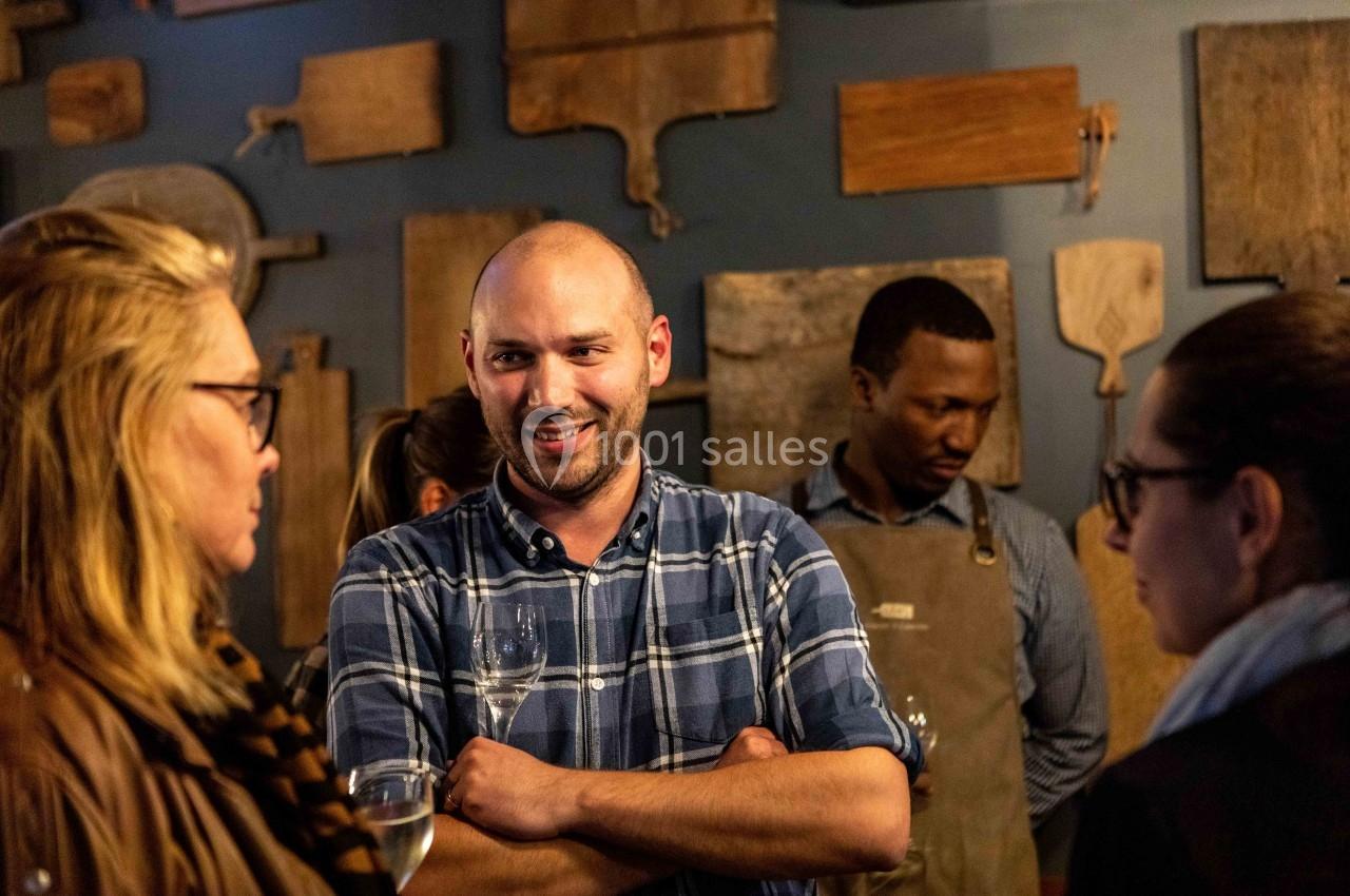 Un homme souriant en chemise à carreaux discute avec des personnes dans un cadre convivial, entouré de planches en bois.