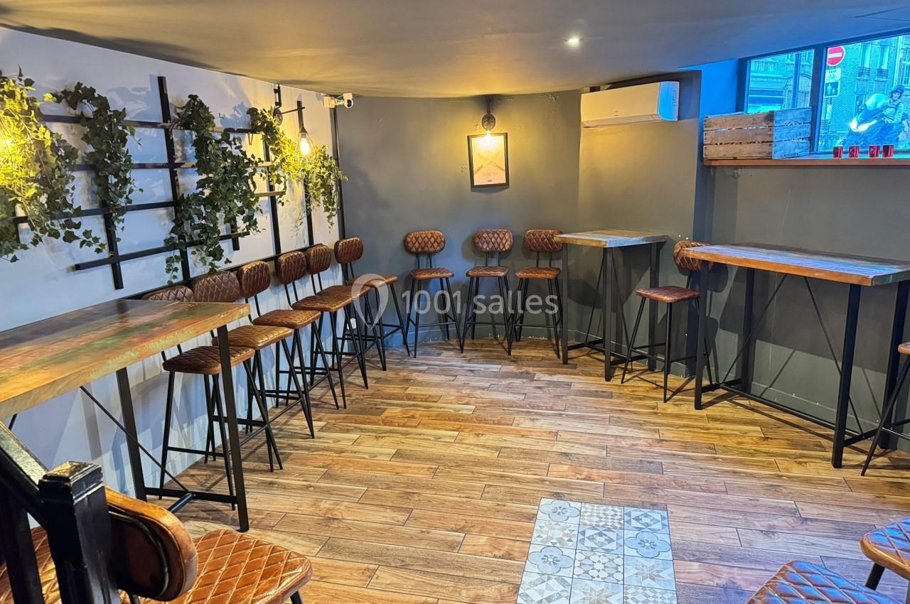 Salle lumineuse avec parquet, tables hautes en bois, chaises en cuir et plantes suspendues sur un mur gris.