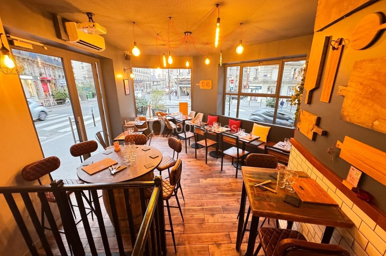 Salle de restaurant chaleureuse avec mobilier en bois, grandes fenêtres et éclairage suspendu.