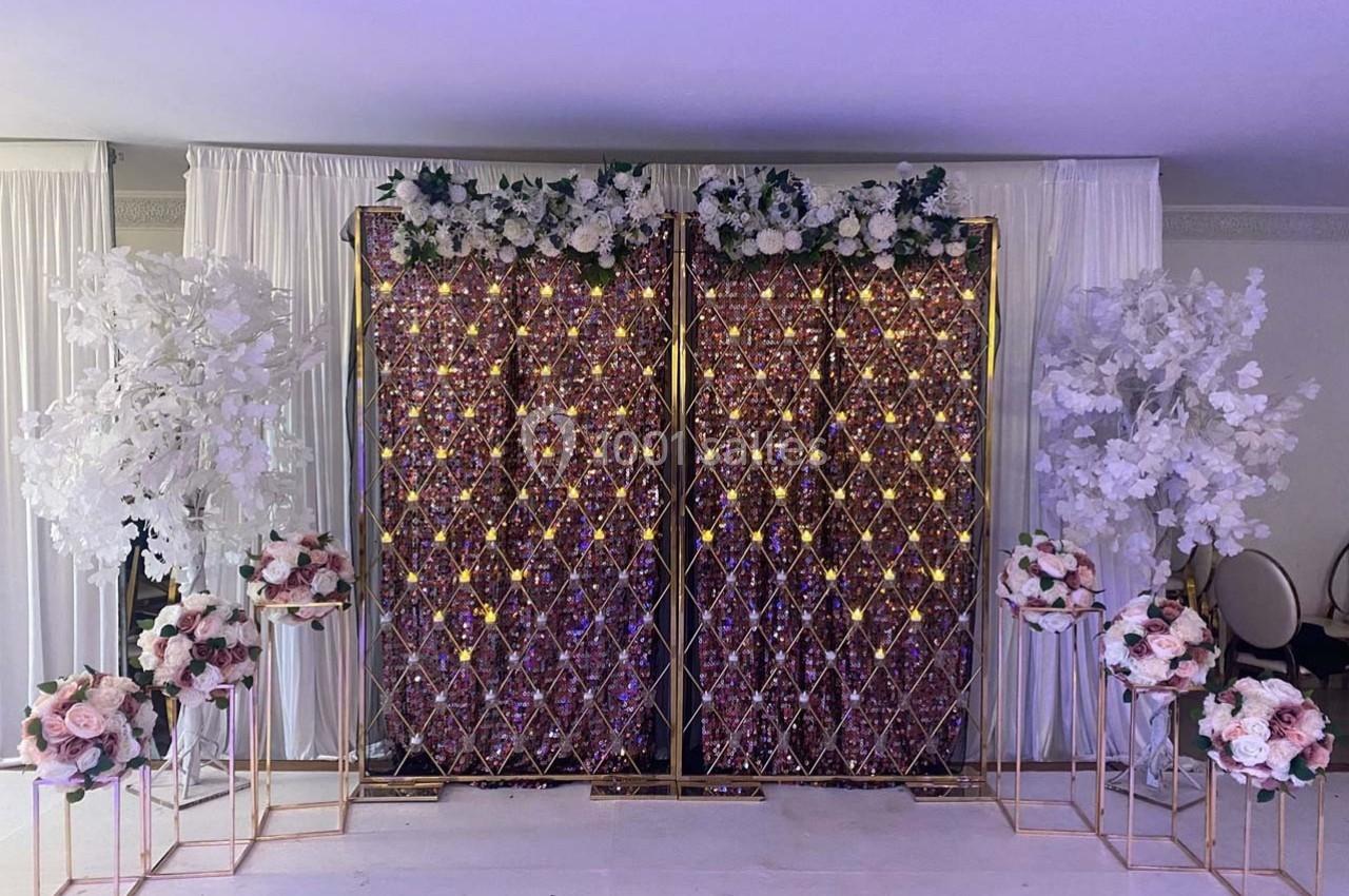 Mur décoratif avec motifs dorés illuminés, fleurs blanches et roses, entouré de rideaux blancs et arbres artificiels.