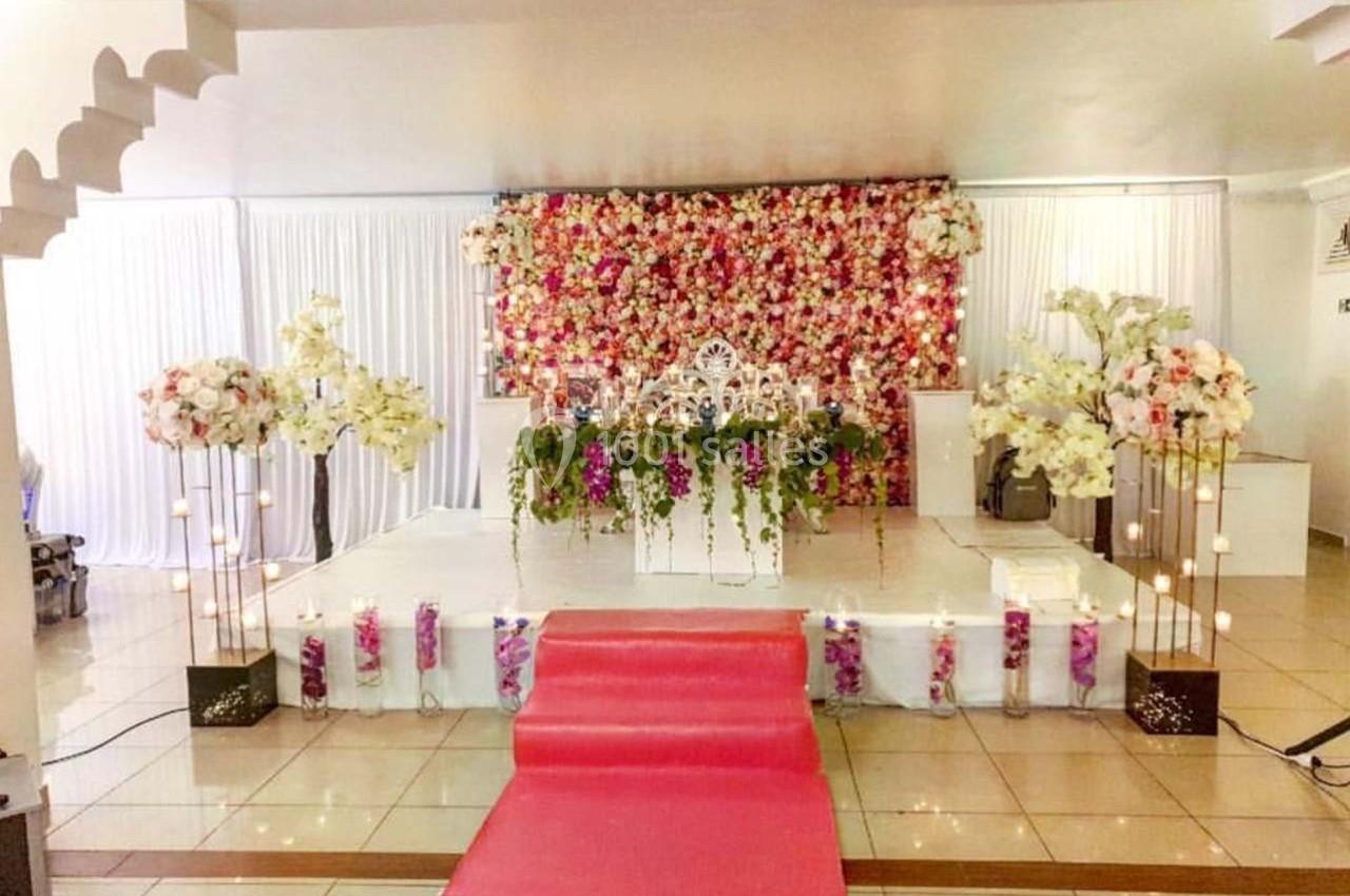 Scène de mariage décorée avec un mur de fleurs colorées, des arrangements floraux et un tapis rouge au centre.