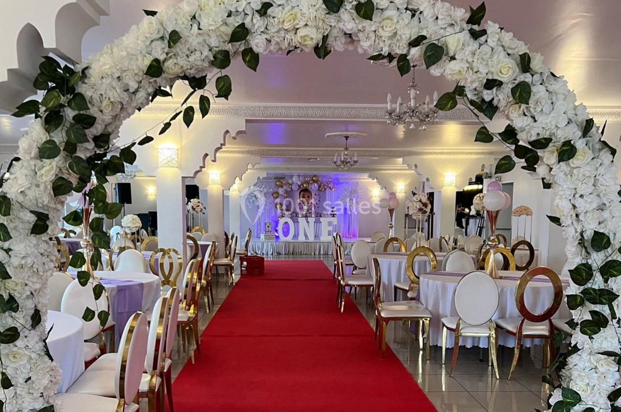 Arche florale blanche décorant une salle de réception avec tables dressées et tapis rouge au centre.