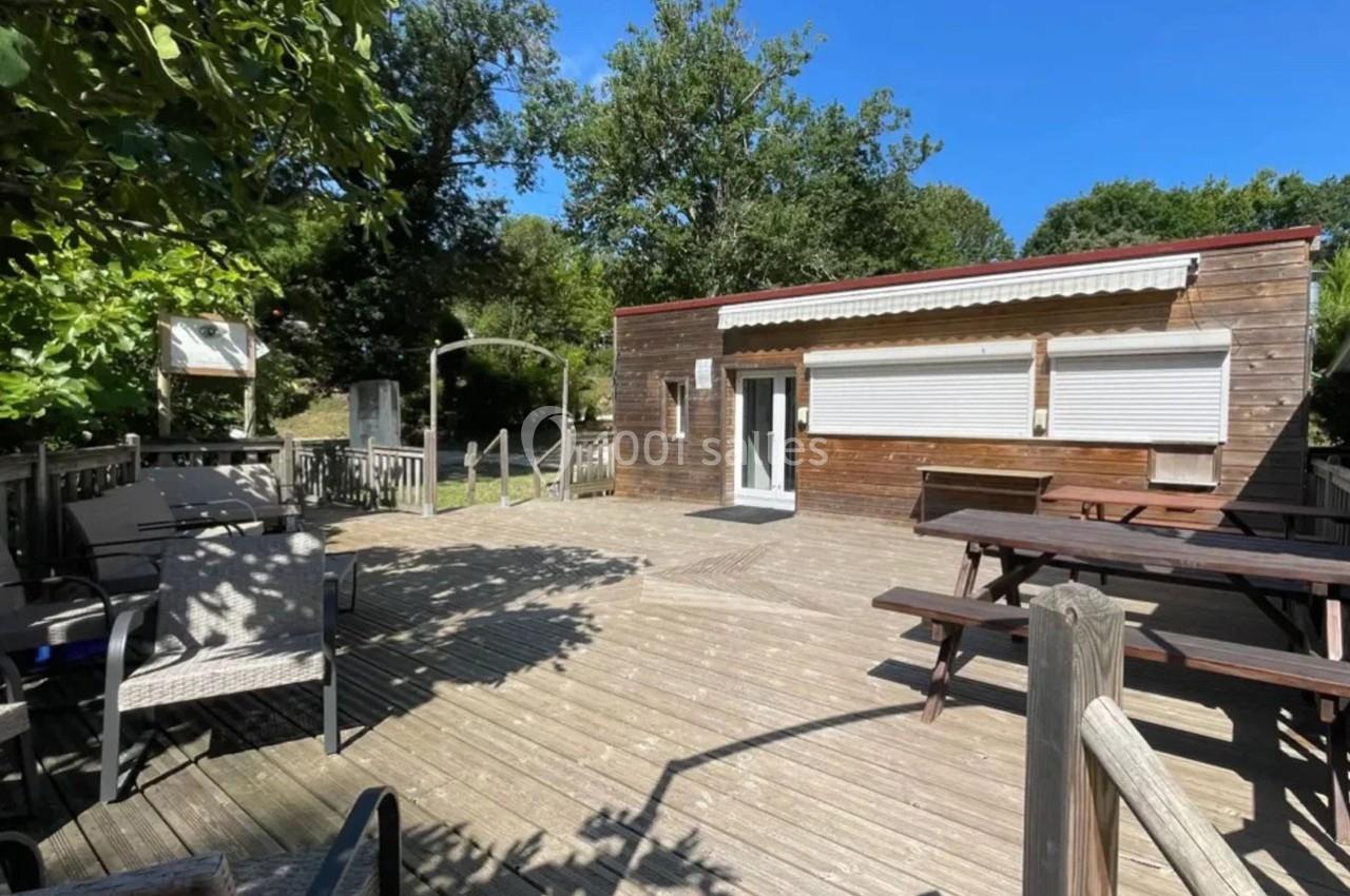 Terrasse en bois avec des tables de pique-nique, des chaises et un bâtiment en bois fermé, entourée de végétation.