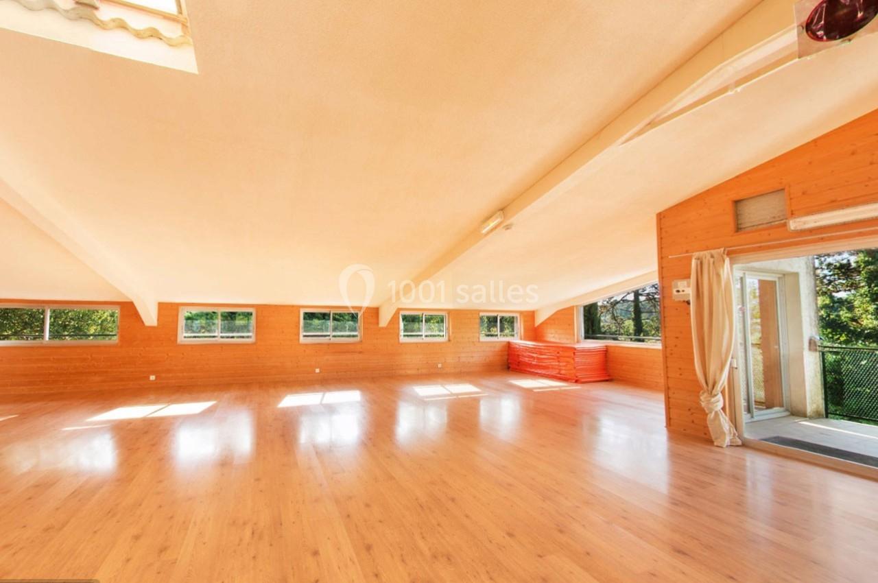 Salle lumineuse avec parquet en bois, murs en lambris, grandes fenêtres et accès à un balcon avec vue sur la verdure.