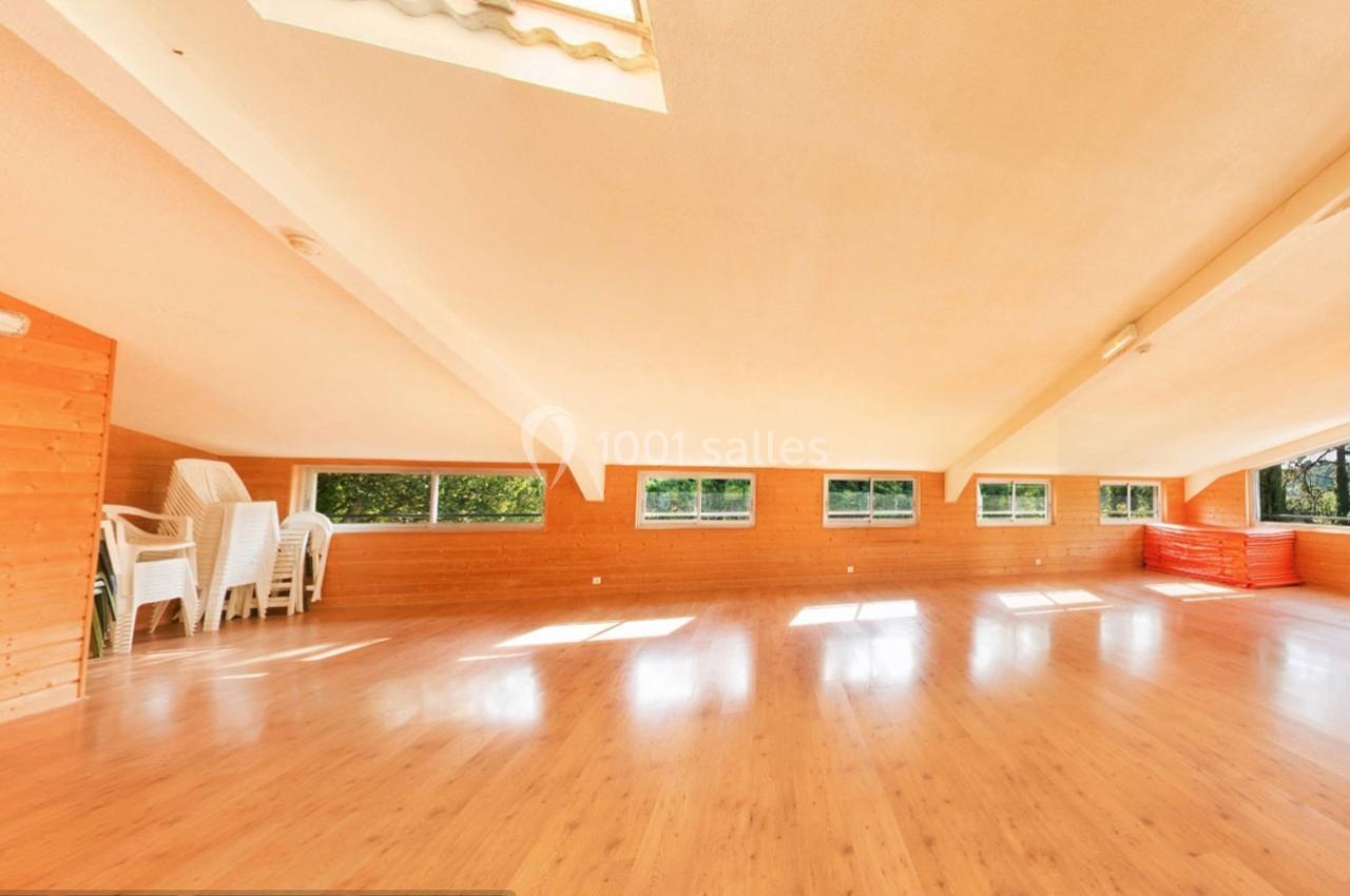 Salle lumineuse avec parquet en bois, murs en bois clair, grandes fenêtres et chaises empilées dans un coin.