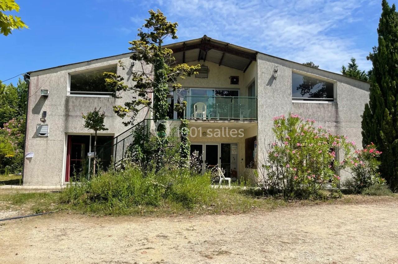 Façade d'une maison moderne à deux étages avec balcon, entourée de végétation et d'un chemin en terre.