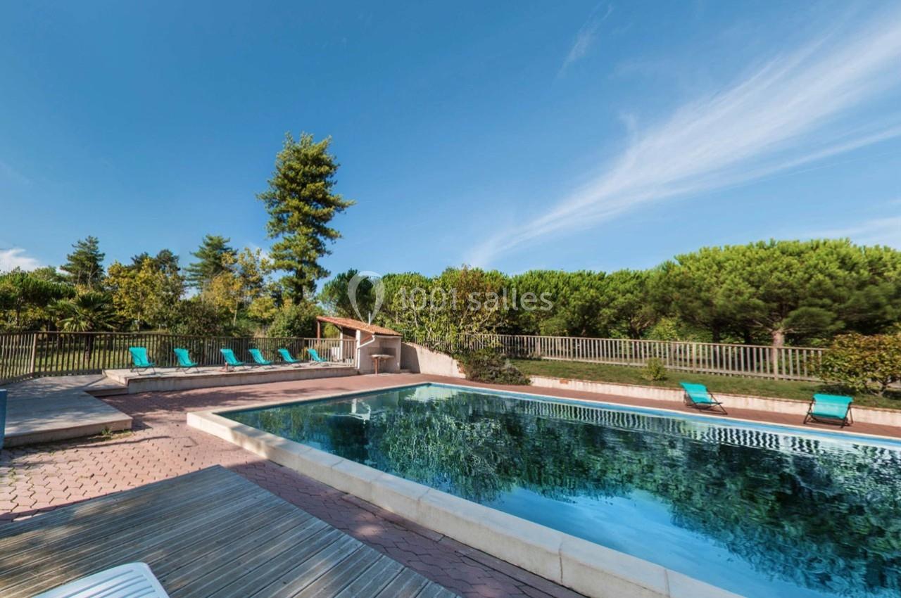 Piscine extérieure entourée de chaises longues, avec végétation et ciel bleu en arrière-plan.