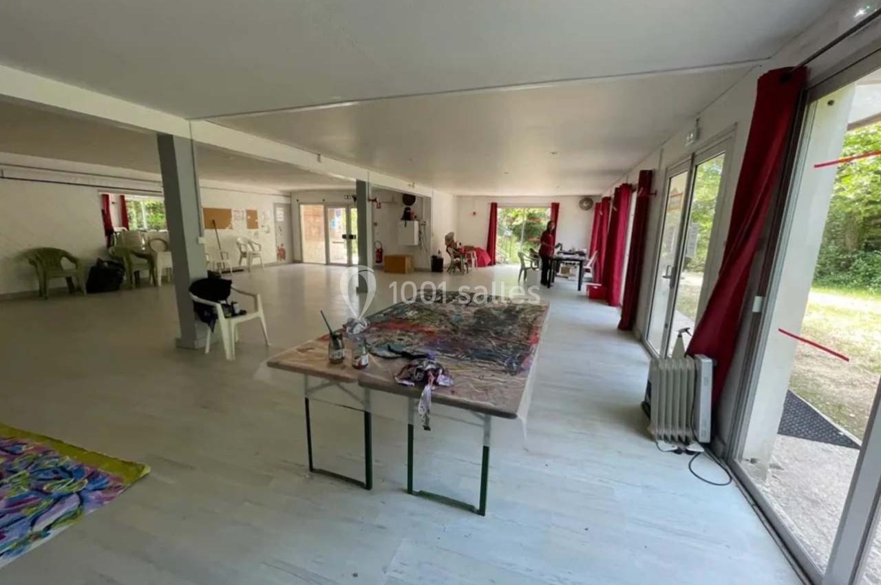 Salle lumineuse avec de grandes baies vitrées, tables de travail, chaises et matériel artistique disposés.