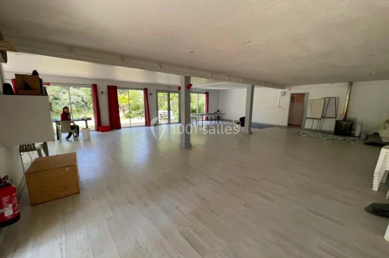 Salle spacieuse avec parquet clair, grandes baies vitrées donnant sur un espace vert, quelques meubles et une personne…