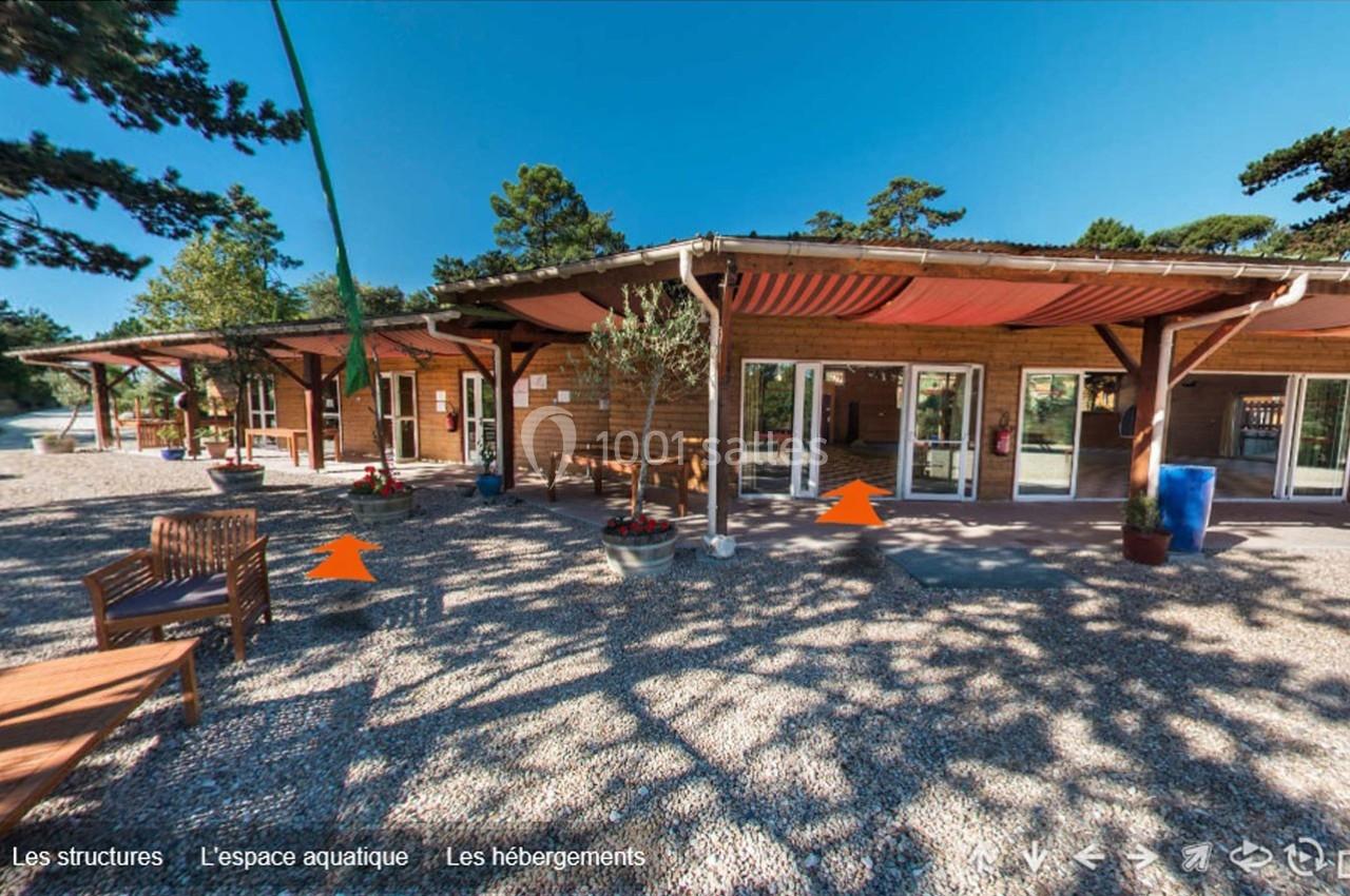 Entrée d'un bâtiment en bois avec terrasse ombragée, entourée de végétation et de mobilier extérieur.