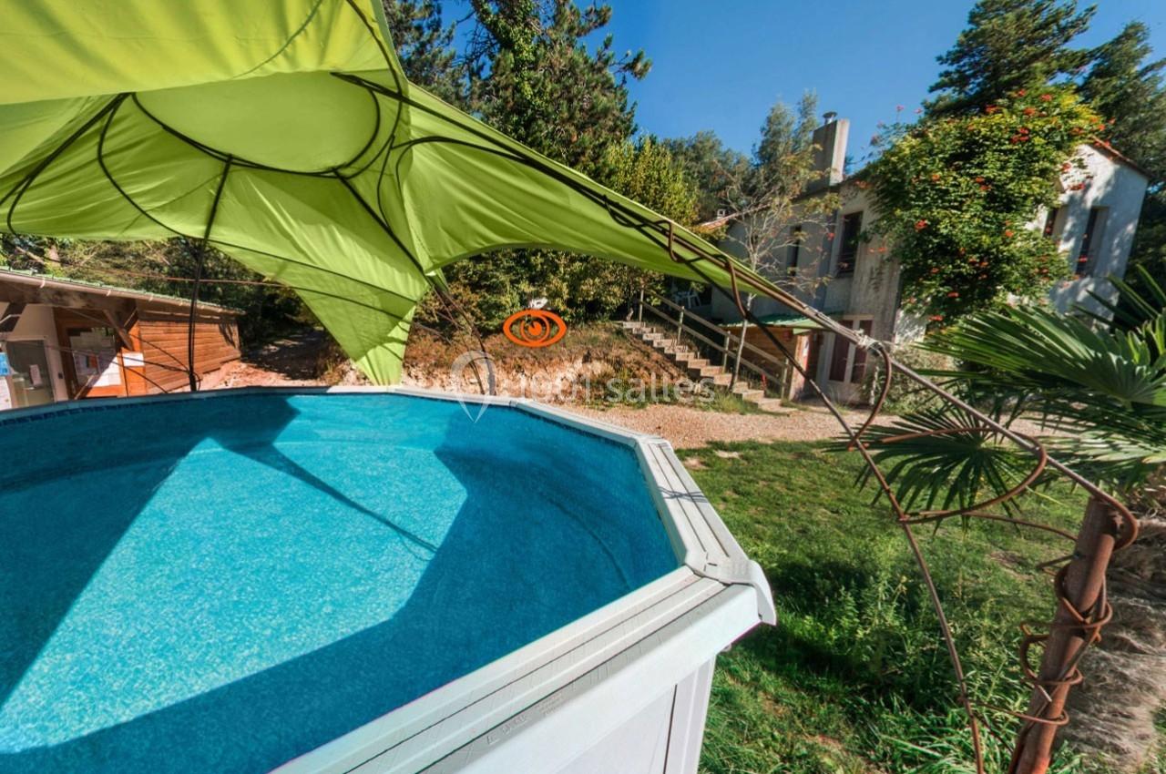 Piscine hors-sol entourée de verdure, avec une maison en arrière-plan et une voile d'ombrage verte.