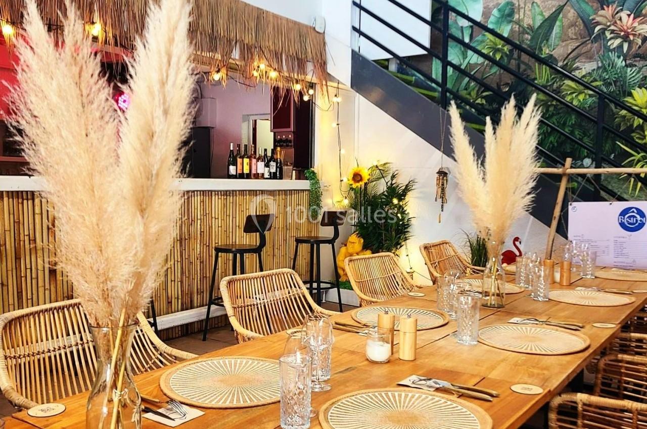 Salle décorée avec des tables en bois, chaises en rotin, vases de pampas et un bar en bambou en arrière-plan.