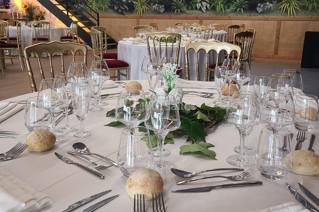 Table dressée pour un repas, avec nappes blanches, couverts, verres, petits pains et décoration végétale en centre.