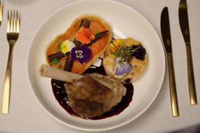 Assiette gastronomique avec un jarret d'agneau, purée de patate douce, gratin et fleurs comestibles, sur une nappe blanche.