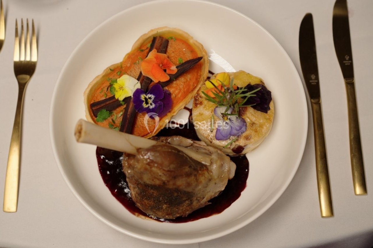 Assiette gastronomique avec un jarret d'agneau, purée de patate douce, gratin et fleurs comestibles, sur une nappe blanche.