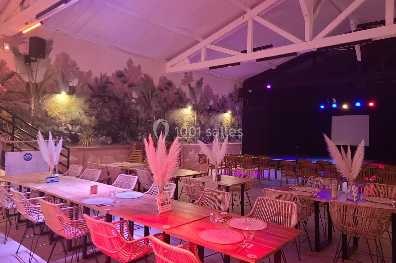 Salle décorée avec des tables en bois, chaises en rotin, pampas en centre de table et éclairage coloré.