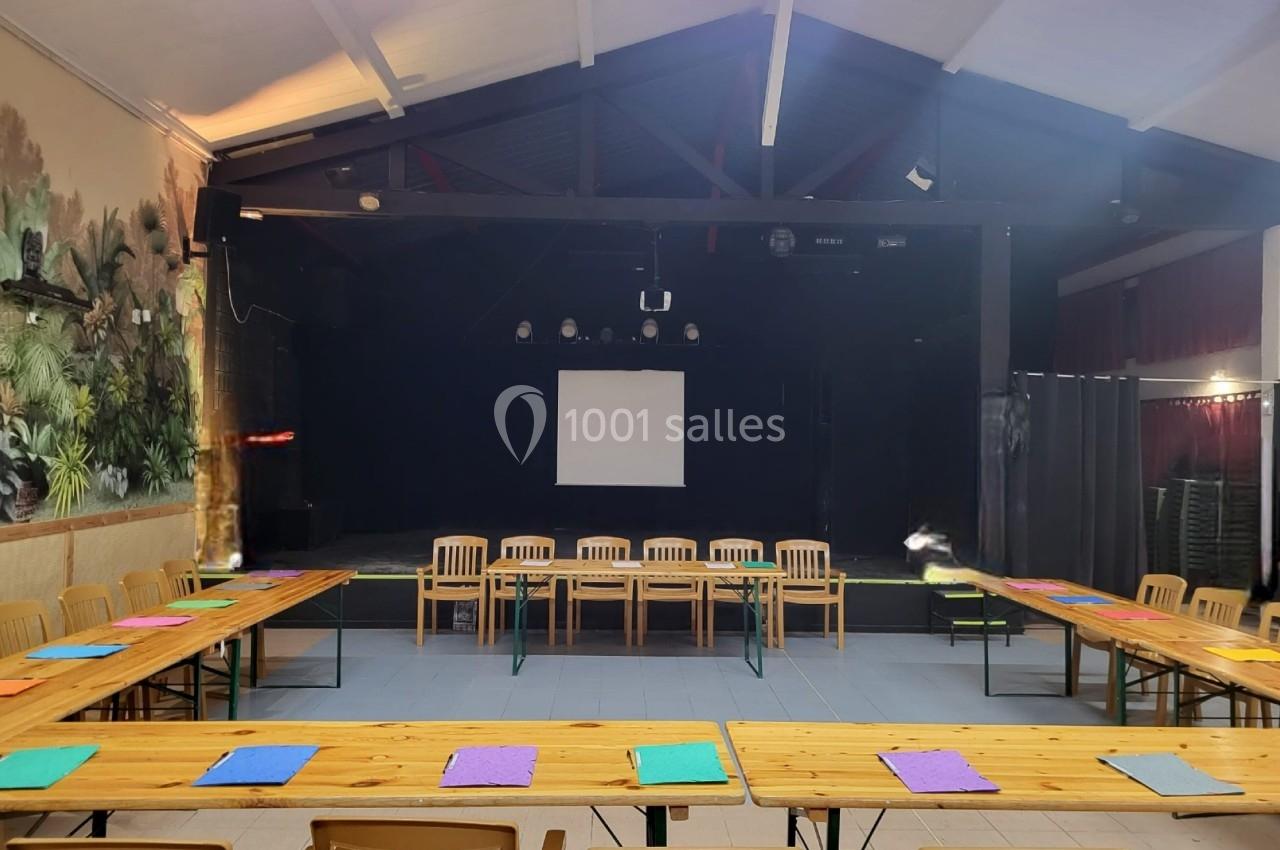 Salle avec une scène, des tables en U disposées avec des feuilles colorées et des chaises en bois, éclairage intérieur.