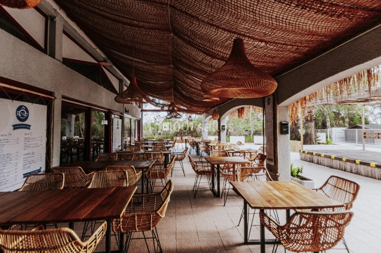 Terrasse couverte avec tables en bois et chaises en rotin, décorée de luminaires suspendus et d'un toit en fibres naturelles.