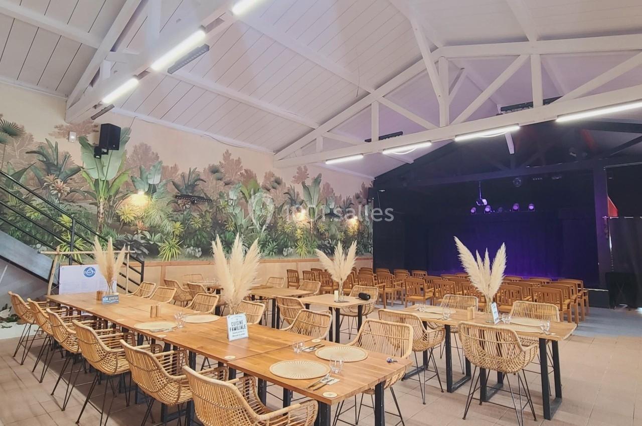 Salle lumineuse avec tables en bois, chaises en rotin, décor mural tropical et scène au fond.
