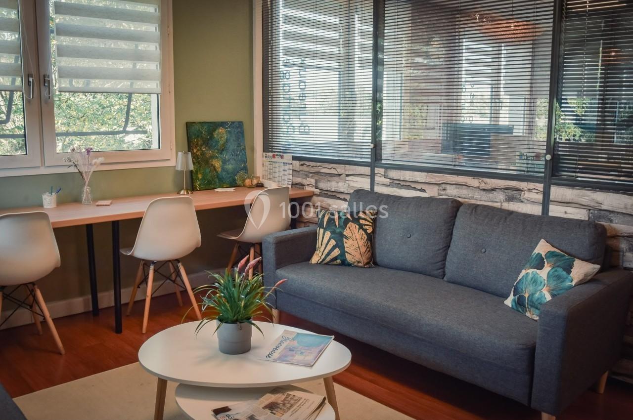 Salon lumineux avec canapé gris, table basse, plantes, et espace de travail avec chaises blanches près d'une fenêtre.