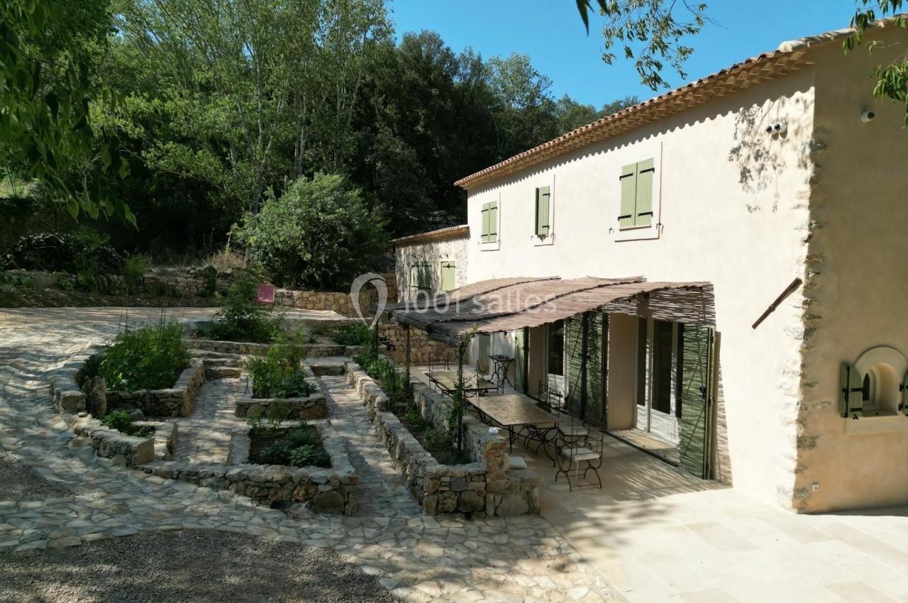 Maison en pierre avec volets verts, terrasse ombragée et jardin aménagé, entourée de végétation et d'une allée pavée.