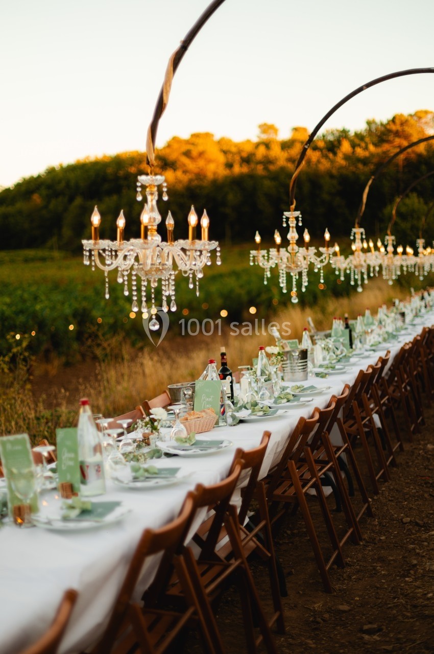 Table dressée en extérieur avec nappes blanches, chandeliers suspendus et décor champêtre au coucher du soleil.
