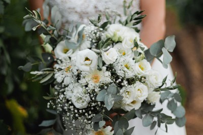 Un couple en tenue de mariage marche main dans la main dans un vignoble, la mariée levant un bouquet.