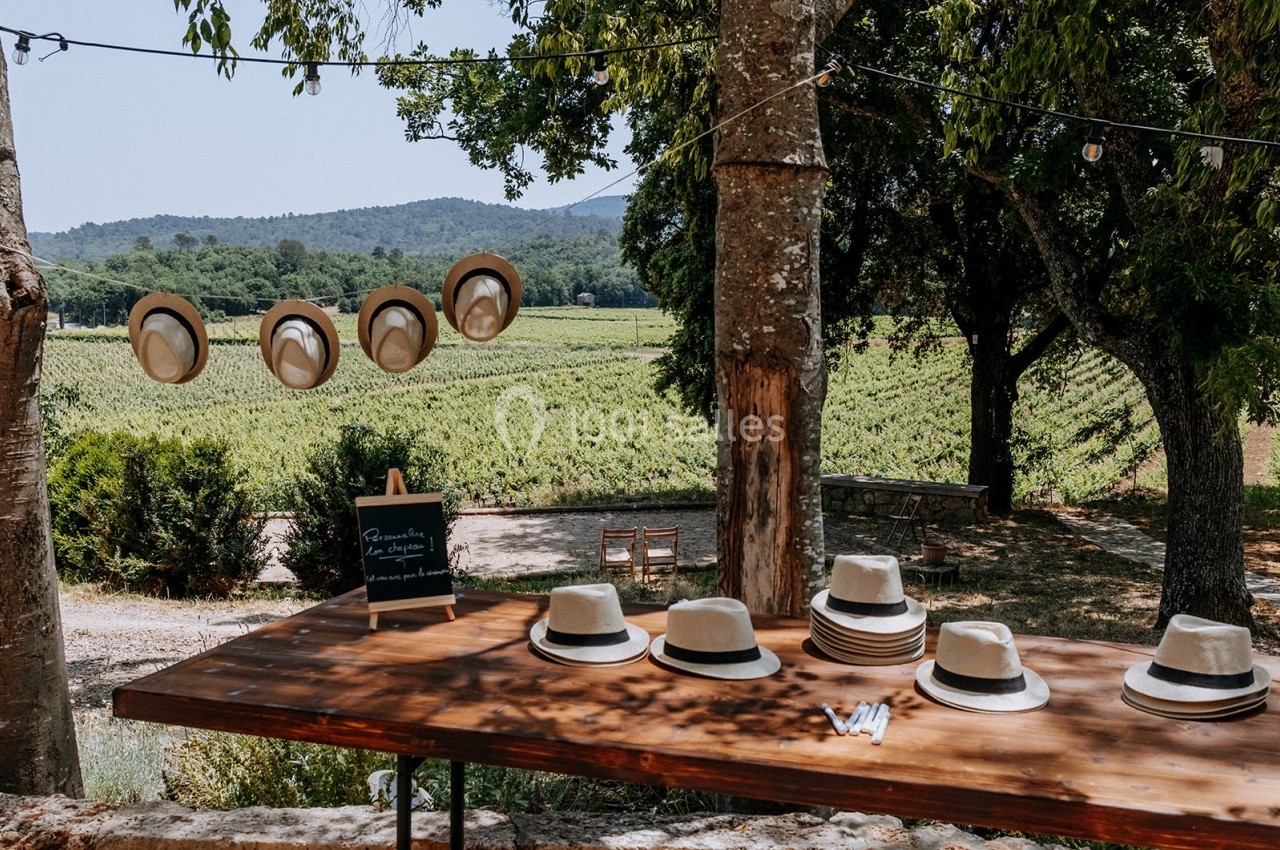 Chapeaux en paille disposés sur une table en bois, avec d'autres suspendus, dans un cadre champêtre près de vignes.