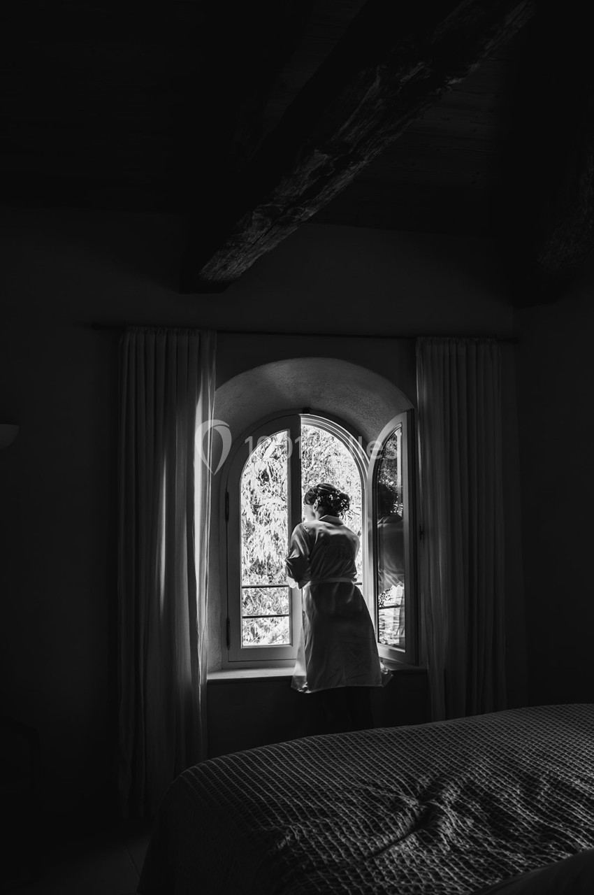 Une femme en robe de chambre regarde par une fenêtre cintrée, éclairée par la lumière naturelle.