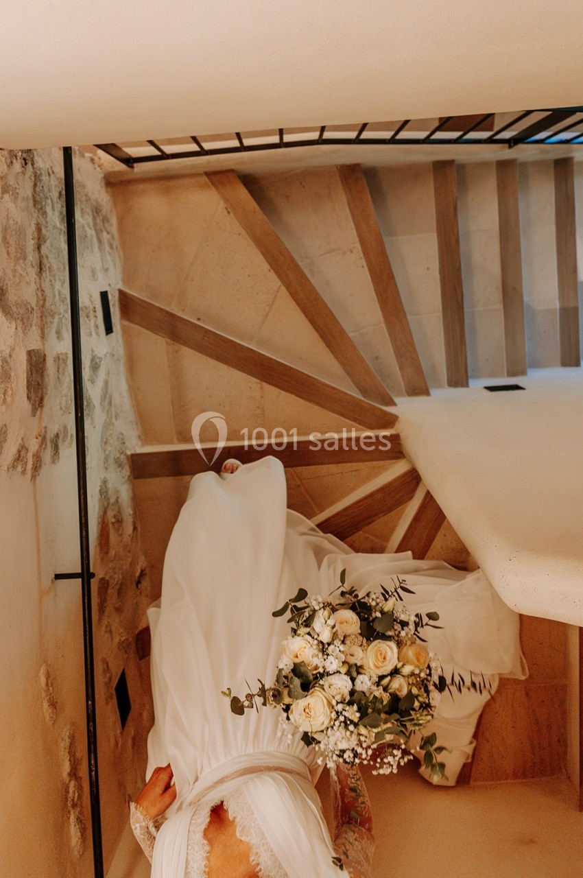 Une mariée en robe blanche descend un escalier en colimaçon, tenant un bouquet de fleurs blanches et jaunes.