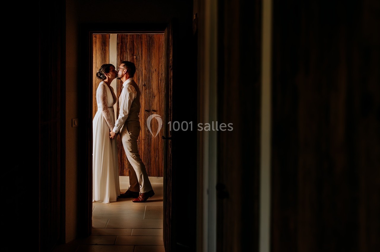 Un couple en tenue de mariage se tient par la main et s'embrasse dans l'encadrement d'une porte en bois.