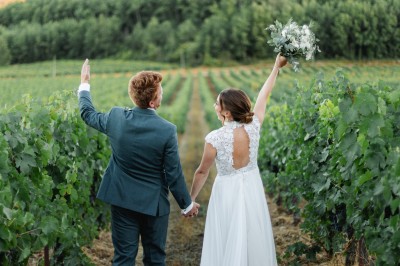 Un couple en tenue de mariage marche main dans la main dans un vignoble, la mariée levant un bouquet.