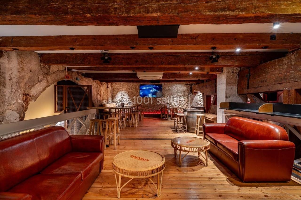 Salle avec murs en pierre, poutres apparentes, canapés en cuir rouge, tables en bois et écran mural allumé.